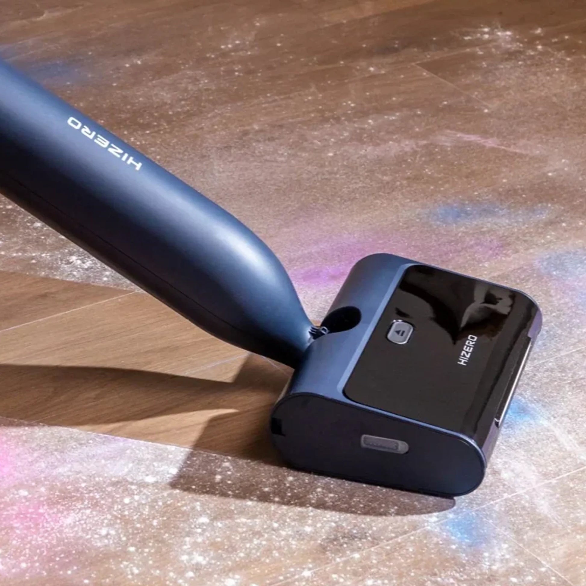 future tech gadgets Canada. smart tech store Canada. HiZero F600 All-in-One Bionic Floor Cleaner. Gallery image 2. The vacuum cleaner is positioned diagonally in the frame, showcasing its sleek design. The body is a deep navy blue with a glossy black base that features a visible power button and the brand name "HIZERO." The vacuum is resting on a wooden floor, with scattered colorful dust and debris around it, illuminated by soft, natural lighting that highlights the surface textures.