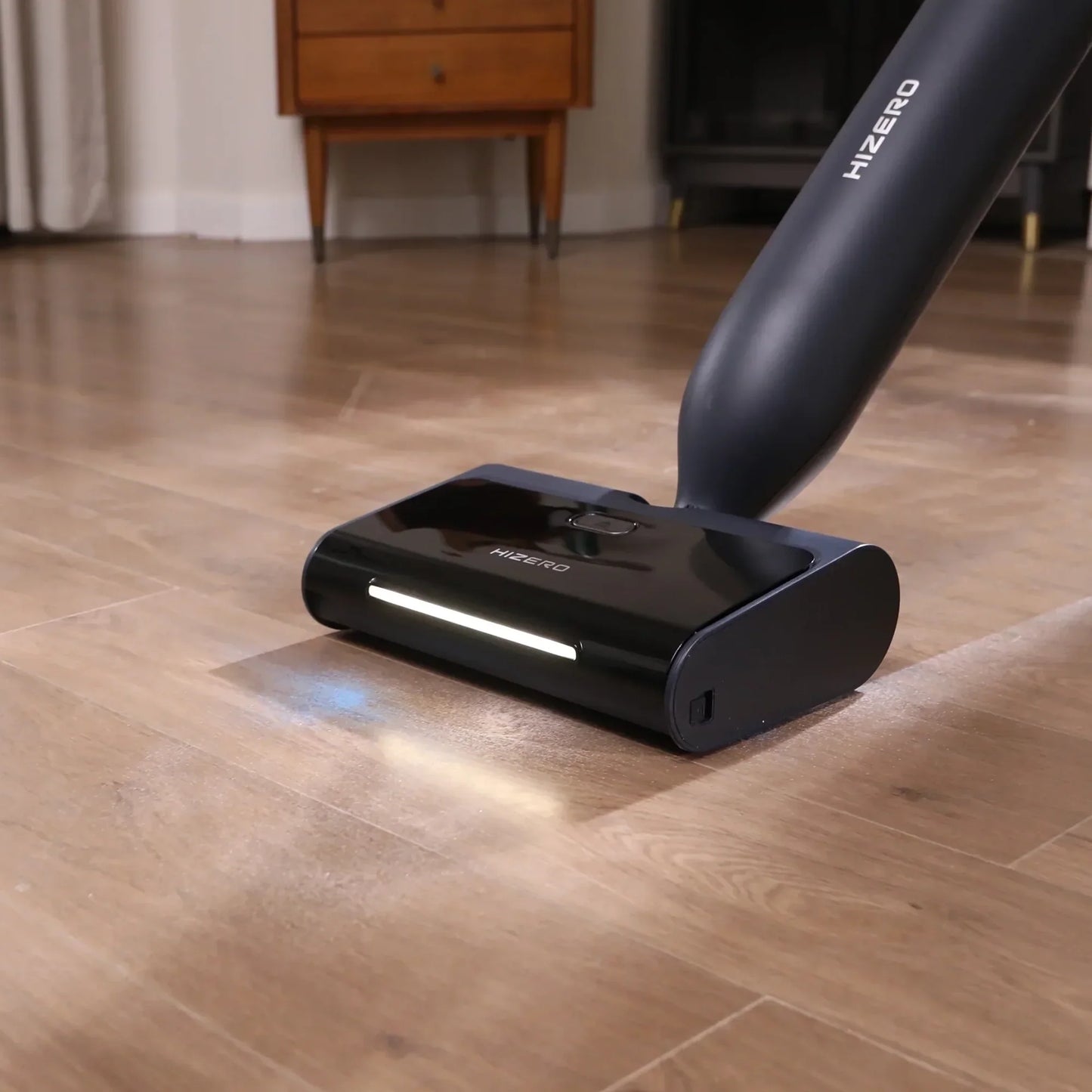 future tech gadgets Canada. smart tech store Canada. HiZero F600 All-in-One Bionic Floor Cleaner. Gallery image 3. The vacuum cleaner is positioned at an angle on a wooden floor, with its cleaning head prominently in the foreground. The sleek, glossy black body features a white LED light strip along the front, illuminating the surface below. The handle extends upward, showcasing the brand name "HIZERO" printed in white on the side.