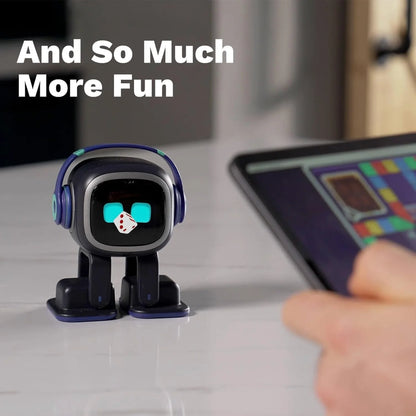 smart tech store Canada. premium tech accessories Canada. Living AI EMO AI Desktop Pet Companion Robot. Gallery image 7. A small, cartoonish robot figure stands on a light-colored surface, centered in the frame. The robot features a square head with large, expressive eyes and is wearing blue headphones. It has a playful design, with a small white die placed on its face. In the background, a hand is reaching towards a tablet displaying a colorful game interface, suggesting interaction.