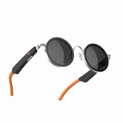 future tech gadgets Canada. smart tech store Canada. Techtron Oracle Smart Sunglasses. Gallery image 6. The sunglasses are displayed at an angle, showcasing their round black lenses and sleek design. The frame features a shiny silver finish with a contrasting matte black section on the arms, which includes a small orange button and four status LED indicators. The tips of the arms are a vibrant orange, adding a pop of color.