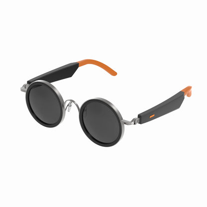 future tech gadgets Canada. smart tech store Canada. Techtron Oracle Smart Sunglasses. Gallery image 8. Round sunglasses are prominently displayed at an angle, showcasing their sleek design. The lenses are dark and circular, framed in shiny silver metal. The arms of the sunglasses feature a matte black finish with vibrant orange accents at the tips. The background is a seamless white, providing a clean contrast that highlights the sunglasses' modern aesthetic.
