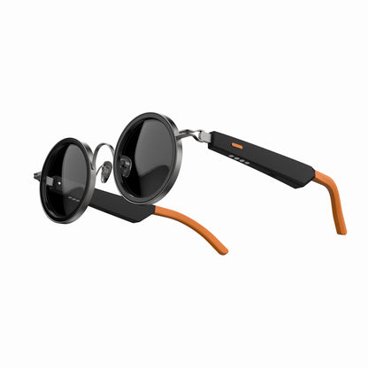 future tech gadgets Canada. smart tech store Canada. Techtron Oracle Smart Sunglasses. Gallery image 2. Round sunglasses are displayed at a slight angle, showcasing their glossy black lenses and sleek metallic frame. The arms of the glasses feature a matte black finish with an orange accent on the tips. The background is a seamless white, emphasizing the product's design. Soft lighting highlights the reflective surfaces of the lenses and frame, creating subtle shadows.