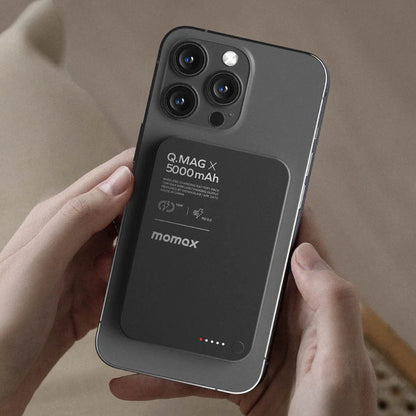 future tech gadgets Canada. premium tech accessories Canada. Momax Q.Mag X 5000mAh Wireless Battery Pack. Gallery image 10. A smartphone is held in two hands, showcasing a sleek, matte black wireless charging battery pack attached to its back. The battery pack features the text "Q.MAG X 5000 mAh" prominently displayed, along with icons indicating charging capabilities. The background is a soft, neutral color, enhancing the product's modern look.