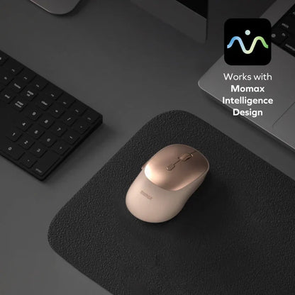 future tech gadgets Canada. smart tech store Canada. Momax 1-Folio A.I. Mouse. Gallery image 3. A sleek, light gold and white wireless mouse is positioned at an angle on a textured black mouse pad, centered in the frame. To the left, a compact black keyboard is partially visible, while a silver laptop sits in the background, slightly blurred. The lighting is soft, creating gentle shadows and highlights on the mouse's glossy surface.