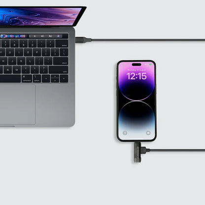 future tech gadgets Canada. premium tech accessories Canada. Momax 1-Link Flow Duo 2-in-1 Braided Cable 1.5M. Gallery image 5. A sleek silver laptop is positioned on the left side of the frame, partially open to reveal a black keyboard and a colorful screen displaying abstract graphics. To the right, a black smartphone is upright, showing a time display of 12:15 against a gradient background. A dark charging cable connects the phone to the laptop, lying flat against a light grey surface.