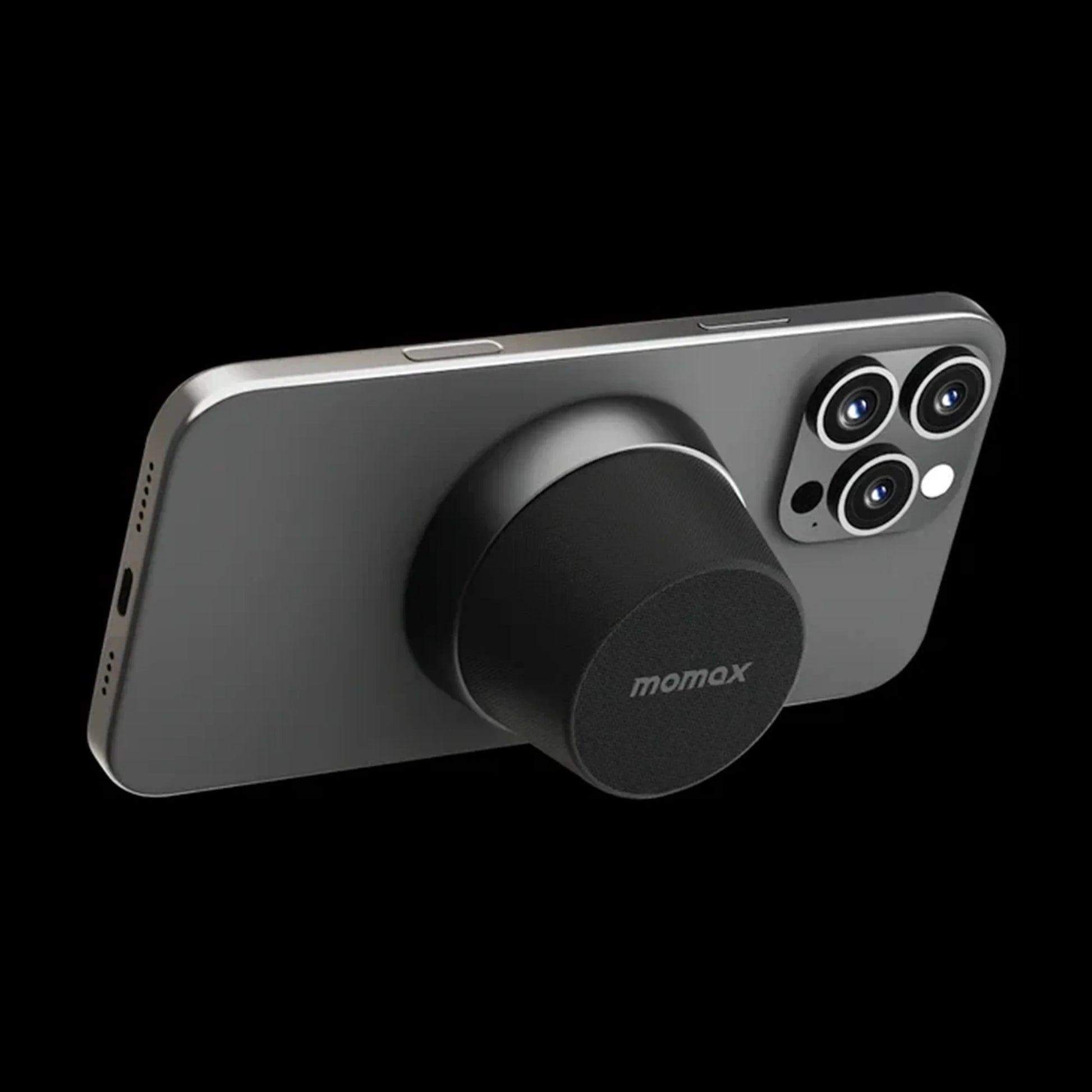 future tech gadgets Canada. premium tech accessories Canada. Momax 1-Vibe Go Magnetic Wireless Speaker. Gallery image 9. The image features a sleek, dark gray smartphone positioned at an angle against a black background. A circular, black accessory with a textured surface is attached to the back of the phone, prominently displaying the "momax" logo. The phone's camera module, which includes three lenses, is visible on the right side.
