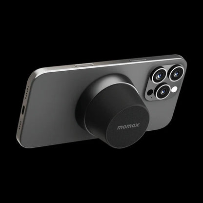 future tech gadgets Canada. premium tech accessories Canada. Momax 1-Vibe Go Magnetic Wireless Speaker. Gallery image 9. The image features a sleek, dark gray smartphone positioned at an angle against a black background. A circular, black accessory with a textured surface is attached to the back of the phone, prominently displaying the "momax" logo. The phone's camera module, which includes three lenses, is visible on the right side.