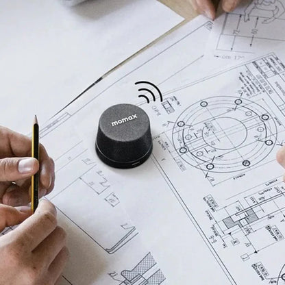 future tech gadgets Canada. premium tech accessories Canada. Momax 1-Vibe Go Magnetic Wireless Speaker. Gallery image 10. A compact, cylindrical speaker with a textured black fabric exterior is positioned on a set of technical drawings spread across a light wooden surface. The speaker features the brand name "momax" printed in white on the front.