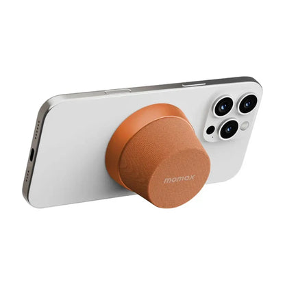future tech gadgets Canada. premium tech accessories Canada. Momax 1-Vibe Go Magnetic Wireless Speaker. Gallery image 12. A smartphone is positioned at an angle, showcasing its sleek white back and three camera lenses on the upper right. Attached to the phone is a round, orange accessory with a textured fabric surface, likely a grip or stand. The background is a seamless white, emphasizing the product's clean design.