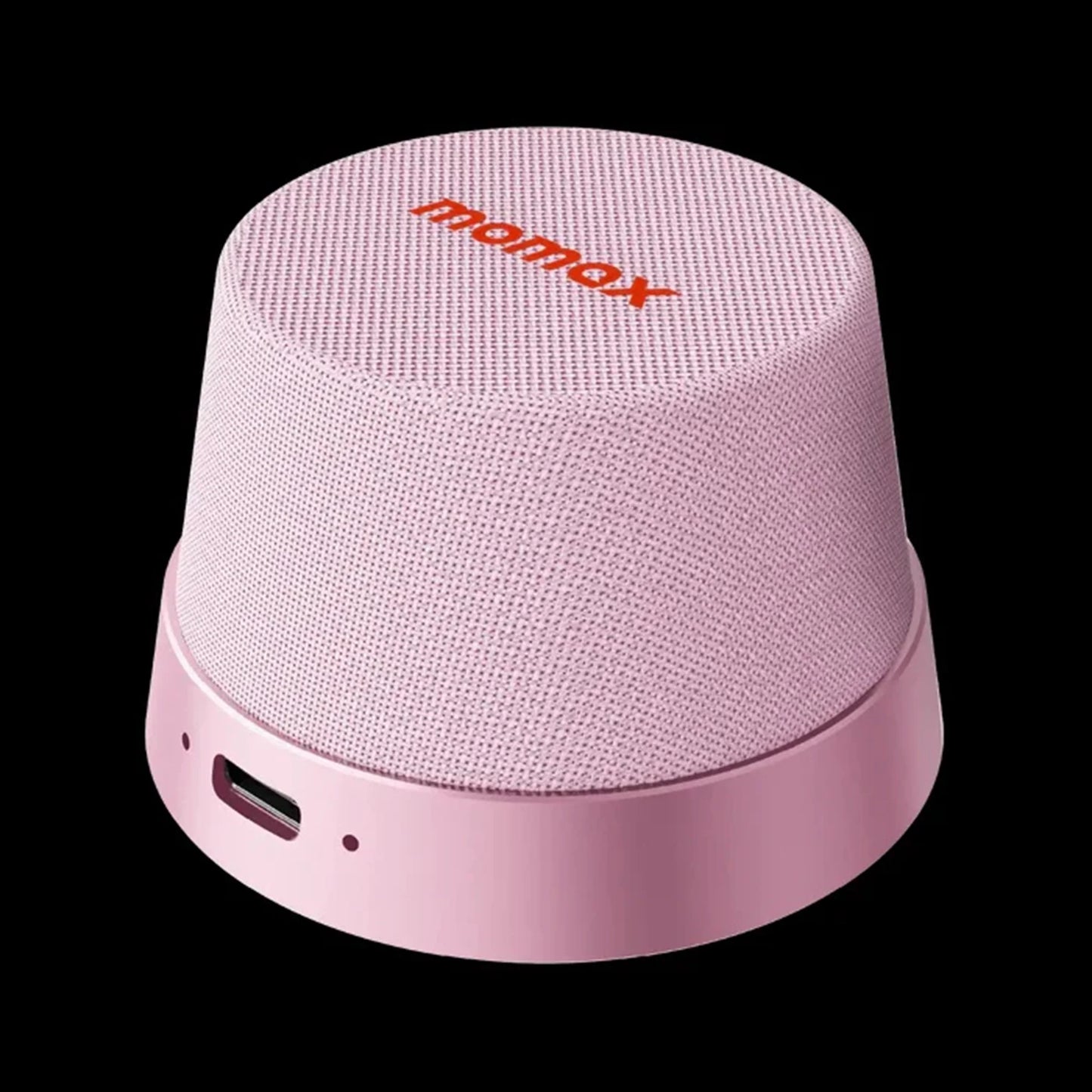 future tech gadgets Canada. premium tech accessories Canada. Momax 1-Vibe Go Magnetic Wireless Speaker. Gallery image 5. The speaker is positioned slightly off-center, showcasing its rounded, compact design. It features a soft pink fabric exterior with a textured finish, and the brand name "momax" is prominently displayed in bright red on the top. The base is a smooth, glossy pink plastic, with a small port and an indicator light visible on the side.