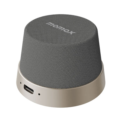 future tech gadgets Canada. premium tech accessories Canada. Momax 1-Vibe Go Magnetic Wireless Speaker. Gallery image 3. The speaker is positioned slightly off-center, showcasing its rounded, cylindrical shape. The top is covered with a textured gray fabric, while the base features a smooth, light gold finish. A small USB-C port is visible on the side, along with a status LED indicator. The background is a seamless white, providing a clean contrast that highlights the speaker's design.