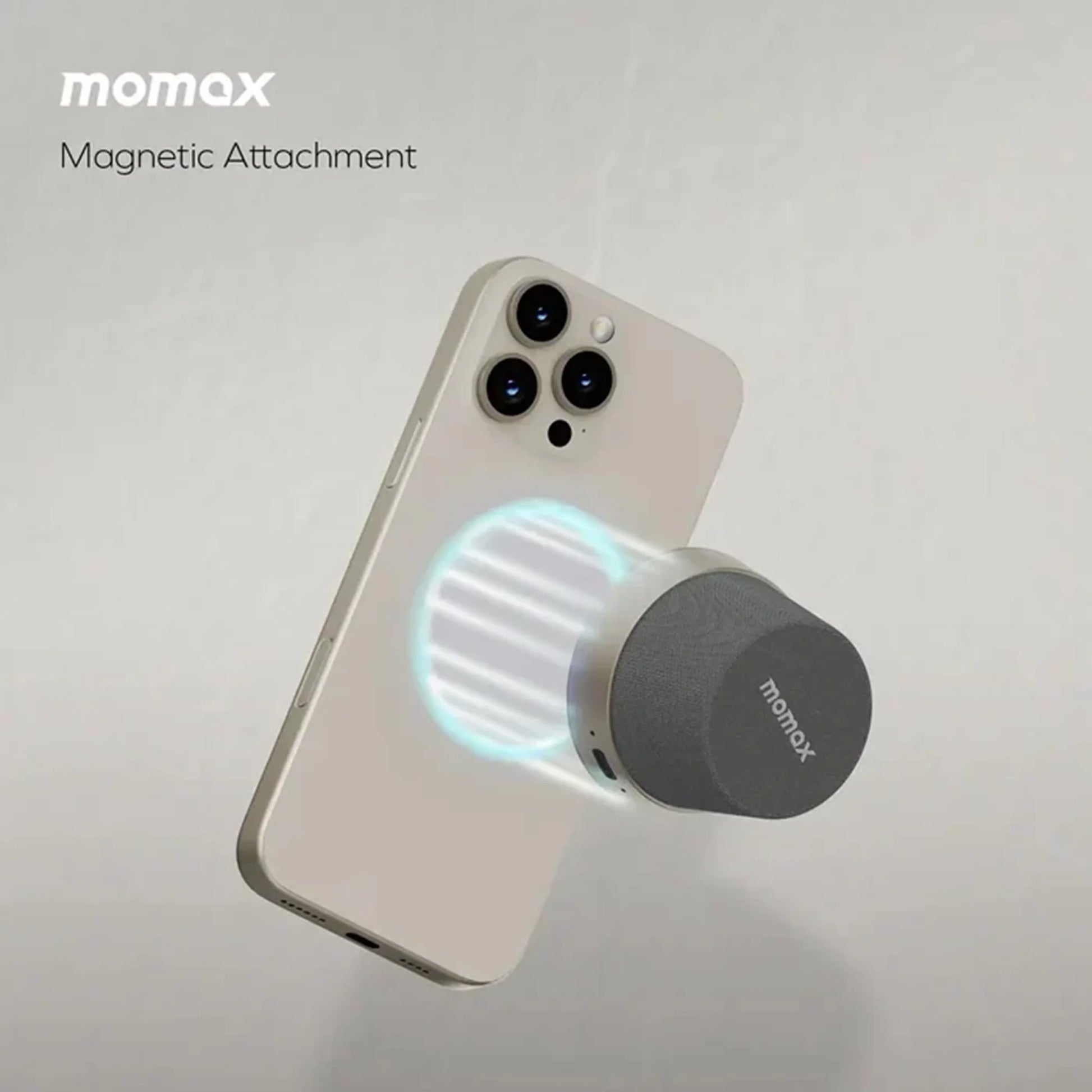future tech gadgets Canada. premium tech accessories Canada. Momax 1-Vibe Go Magnetic Wireless Speaker. Gallery image 17. A light-colored smartphone is positioned at an angle, showcasing its three rear cameras and a magnetic attachment device hovering near its back. The attachment features a cylindrical design with a dark gray fabric texture on top and a smooth metallic base, emitting a soft blue glow around the contact area.