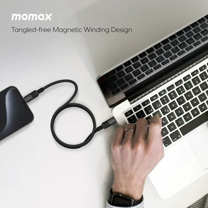 future tech gadgets Canada. smart tech store Canada. Momax Mag.Link 100W Usb-C To Usb-C Magnetic Cable 1M. Gallery image 7. A sleek, black portable charger is positioned to the left of a silver laptop, with a flexible charging cable extending from the charger to the laptop's USB port. The charger features a smooth, matte finish, while the cable is coiled in a way that suggests a tangle-free design. A hand, wearing a black watch, is reaching towards the laptop's keyboard, indicating active use.