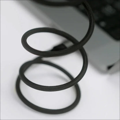 future tech gadgets Canada. premium tech accessories Canada. Momax Mag.Link 66W Usb-A To Usb-C Magnetic Cable 1M. Gallery image 3. A close-up view of a coiled black charging cable is prominently displayed in the foreground, with three loops visible. The cable features a textured surface, suggesting durability. In the background, a portion of a silver laptop is slightly blurred, indicating the cable is connected to it.