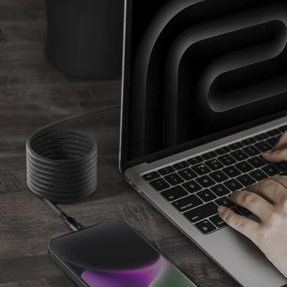 next gen electronics shop. buy electronics online Canada. Momax Mag.Link Usb-C To Lightning Magnetic Cable 2M. Gallery image 3. A sleek, silver laptop is positioned at an angle on a dark wooden surface, with a hand resting on its black keyboard. A coiled, black braided charging cable extends from the laptop to a smartphone lying flat beside it, displaying a colorful screen. The background features a dark, abstract design, enhancing the modern aesthetic.