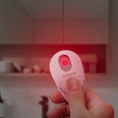 future tech gadgets Canada. smart tech store Canada. Momax Pinguard Camera Scanning Find My Locator. Gallery image 7. A hand holds a small, oval-shaped remote control with a glossy pink finish. The remote features a prominent red laser light activated by a button, which is currently illuminated. The background is softly blurred, revealing a light-colored kitchen setting with shelves and indistinct objects. The lighting is warm and inviting, creating a cozy atmosphere.
