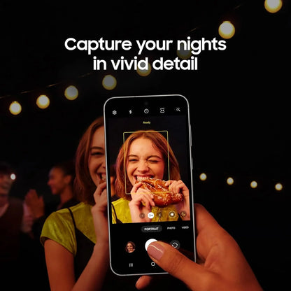 future tech gadgets Canada. smart tech store Canada. Premium Samsung Galaxy S23 FE. Gallery image 9. A smartphone is held in a hand, angled slightly towards the viewer, displaying a camera interface. The screen shows a smiling young woman with medium-length brown hair, holding a pretzel, illuminated by soft, warm lighting from string lights in the background. The background is dark, creating a contrast that highlights the subject.