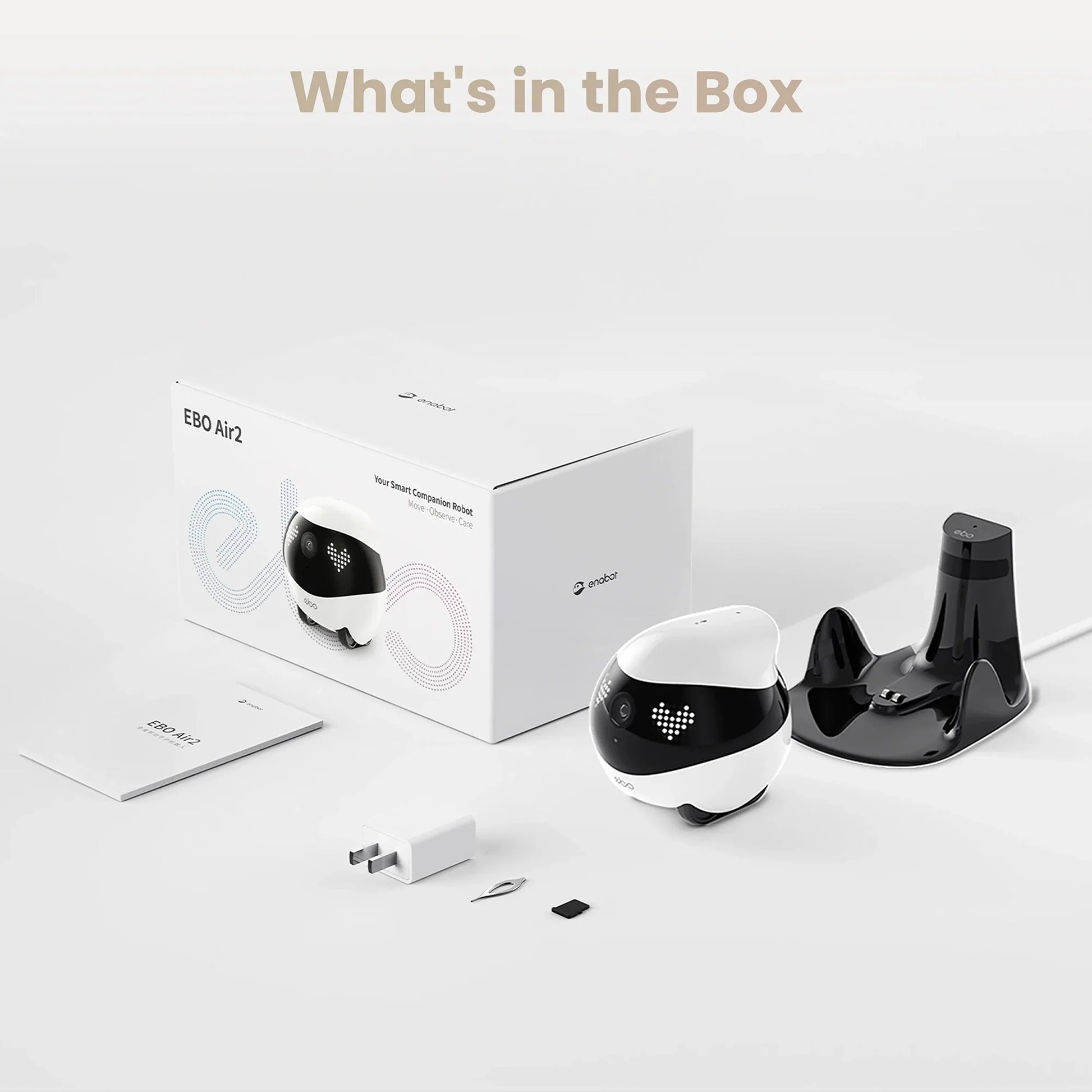 smart tech store Canada. premium tech accessories Canada. Enabot Ebo Air 2 Companion Robot. Gallery image 4. A compact, round robot, primarily white with a black top, is positioned to the left of a white box labeled "EBO Air2" in colorful text. The robot features a small camera and LED lights on its front. To the right, a glossy black charging dock is visible, along with a white power adapter, a small tool, and a memory card.