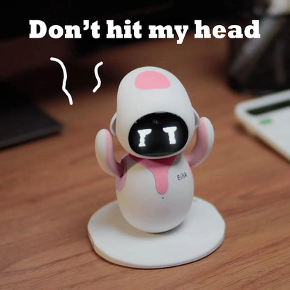 smart tech store Canada. premium tech accessories Canada. Eilik Desktop Companion AI Robot. Gallery image 8. A small, cartoonish robot stands on a circular white base, centered in the frame. The robot has a smooth, glossy white body with pink accents on its arms and head. Its face features a black screen displaying two white eyes and a frowning mouth, conveying a playful expression. The background is a warm wooden surface, with a blurred electronic device partially visible to the right.