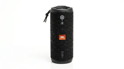future tech gadgets Canada. smart tech store Canada. JBL Flip 7 Portable waterproof and drop-proof speaker. Gallery image 5. The speaker is positioned centrally against a seamless white background, showcasing its cylindrical shape. It features a textured black fabric exterior with visible water droplets, indicating its water-resistant design. The front displays three buttons: a power button, a Bluetooth pairing button, and a volume control, all in a contrasting color.