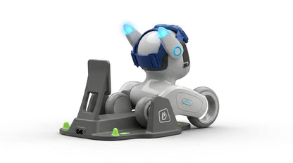 smart tech store Canada. premium tech accessories Canada. Loona AI Pet Robot. Gallery image 8. The image features a small, robotic dog positioned on a gray charging dock, angled slightly to the right. The robot has a glossy white body with blue accents, including a pair of blue headphones and glowing blue lights on its ears. The dock includes green rubber feet for stability and a visible charging port.