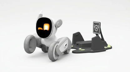 smart tech store Canada. premium tech accessories Canada. Loona AI Pet Robot. Gallery image 10. A small, white and gray robotic toy is positioned slightly to the left of the frame, facing forward. Its round body features a large, black screen displaying two bright, orange eyes that give it a friendly expression. To the right, a black charging dock is visible, with a power symbol icon and green accents.