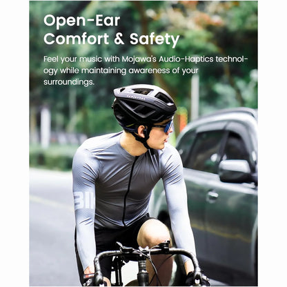 future tech gadgets Canada. smart tech store Canada. Mojawa HaptiFit Terra Head Wearable Sport System. Gallery image 2. A cyclist is positioned slightly to the left of the frame, wearing a fitted, long-sleeve gray jersey with a zippered front and black shorts. He has a black helmet and sunglasses, and is leaning forward on a black bicycle with drop handlebars. The background features a blurred street scene with greenery and a gray vehicle passing by.