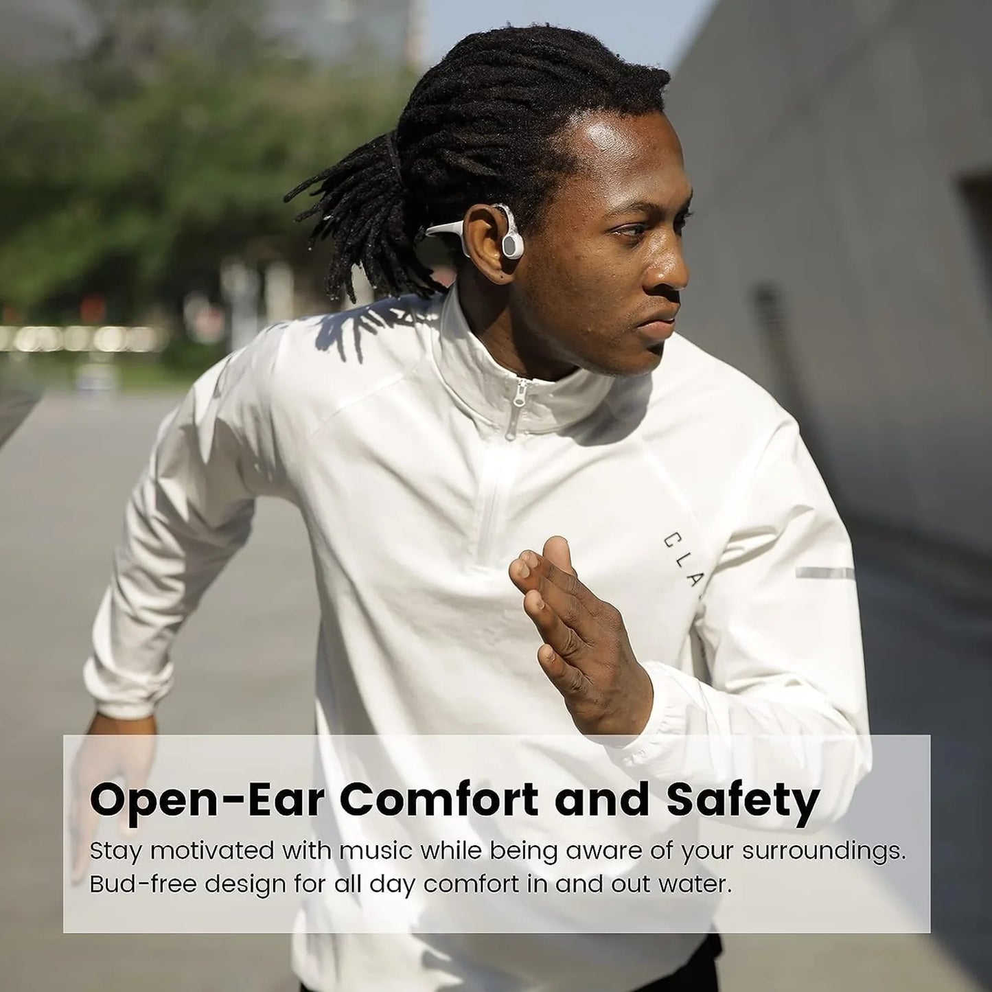 future tech gadgets Canada. smart tech store Canada. Mojawa RUN PLUS Bone Conduction Headphones. Gallery image 2. A young man with dreadlocks is captured mid-run, angled slightly to the right, wearing a light-colored, zip-up jacket. He has a pair of white, open-ear headphones visible in his ears, designed for comfort and safety. The background features a blurred outdoor setting with greenery and a concrete wall, suggesting an active environment.