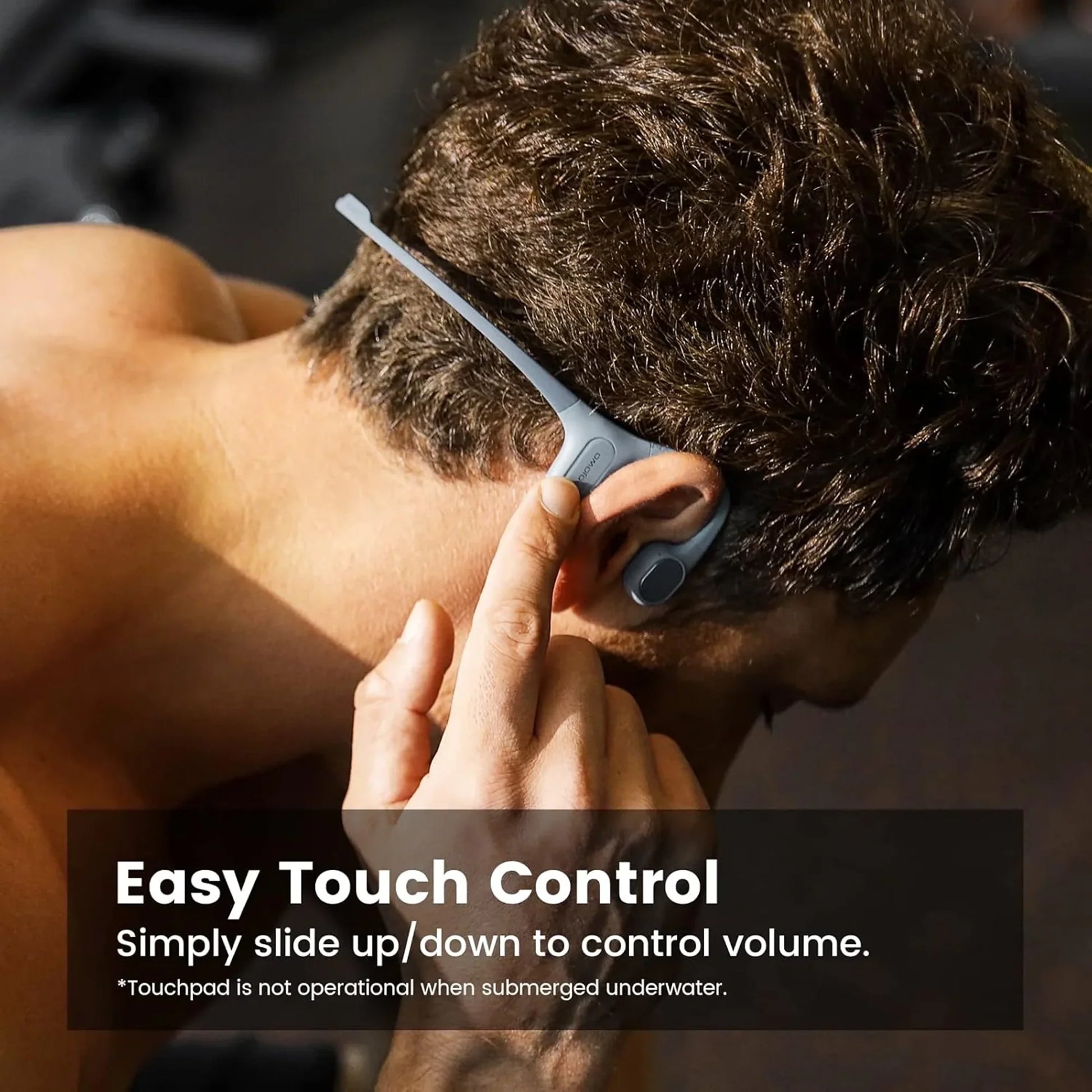 future tech gadgets Canada. smart tech store Canada. Mojawa RUN PLUS Bone Conduction Headphones. Gallery image 5. A person with short, dark hair is shown from the back, adjusting a pair of gray wireless headphones that rest behind their ears. The headphones feature a sleek, curved design with a touch control area highlighted by a small button. The background is softly blurred, suggesting a gym environment with dim lighting that creates gentle highlights on the headphones.