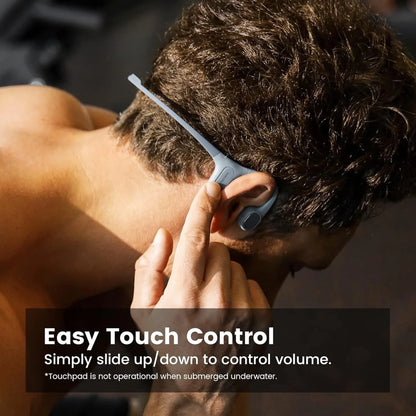 future tech gadgets Canada. smart tech store Canada. Mojawa RUN PLUS Bone Conduction Headphones. Gallery image 5. A person with short, dark hair is shown from the back, adjusting a pair of gray wireless headphones that rest behind their ears. The headphones feature a sleek, curved design with a touch control area highlighted by a small button. The background is softly blurred, suggesting a gym environment with dim lighting that creates gentle highlights on the headphones.